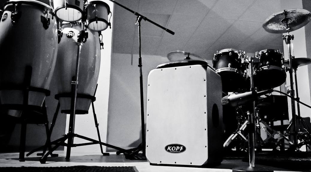 Cajon Setup for Acoustic Gigs – Minimal Stage Layout by Kopf Percussion A clean, minimal acoustic stage setup featuring a handcrafted cajon, two microphones on stands, and a simple wooden stool under soft studio lighting. The scene reflects the balanced, natural tone and simplicity that define Kopf Percussion’s approach to live performance setups.