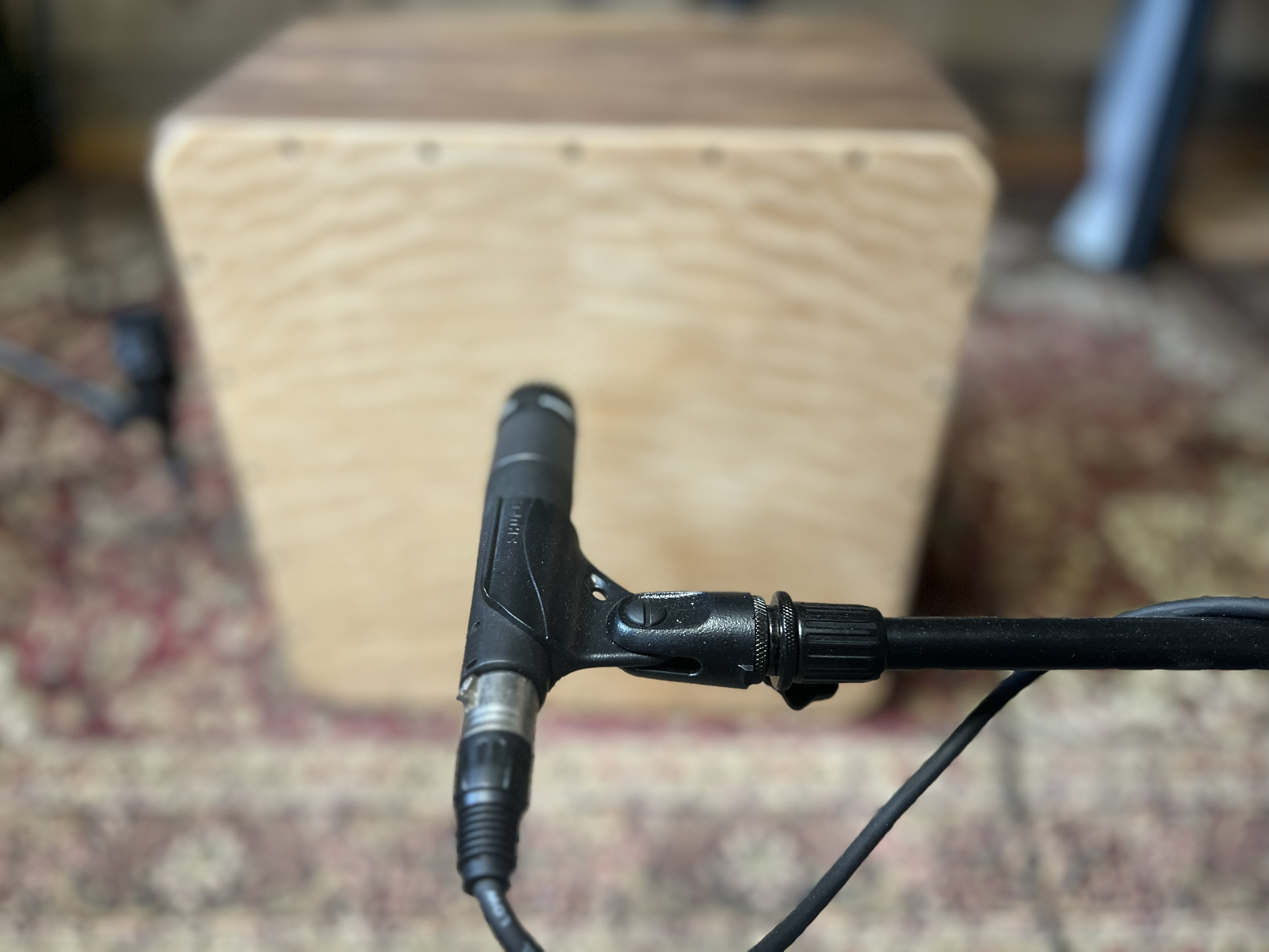 Front Microphone Placement on Cajon – Kopf Percussion A close-up view of a Kopf Percussion cajon with a microphone positioned in front of the faceplate, aimed high between the player’s knees toward the snare area. This placement captures natural tone and articulation while maintaining a balanced, acoustic sound.