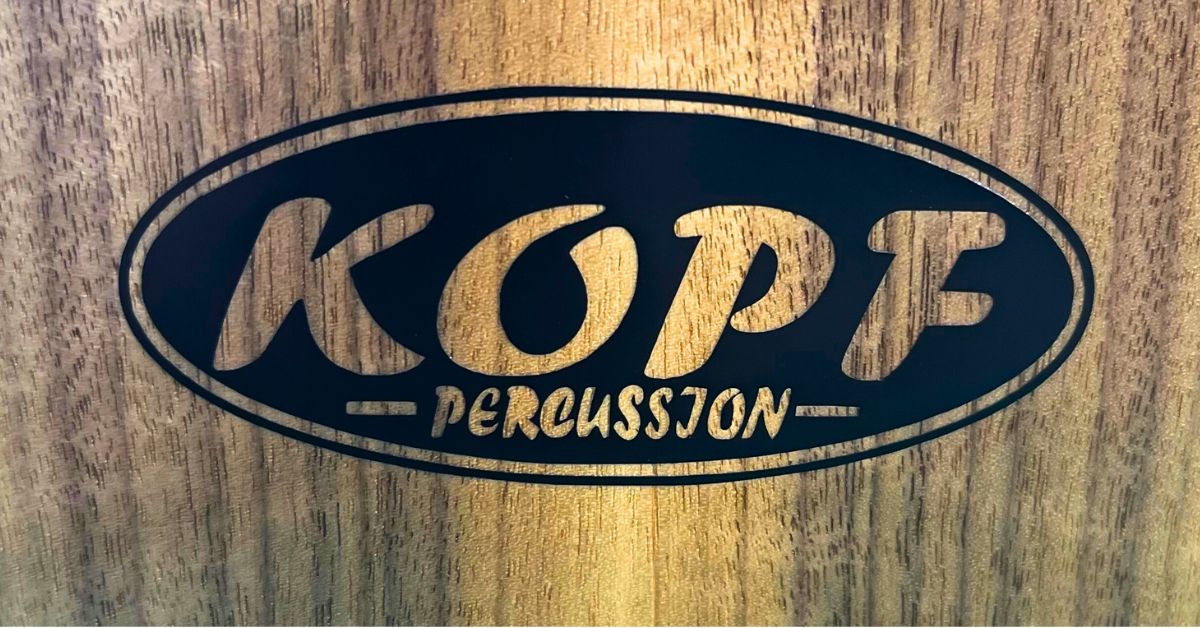 Close-up of the Kopf Percussion maker&rsquo;s mark set into a curly walnut cajon faceplate, representing authorship and responsibility in handcrafted percussion.